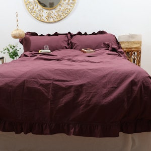 May include: A burgundy bed with a ruffled duvet cover and two matching pillowcases. The bed is made with white sheets and has a white bed skirt.