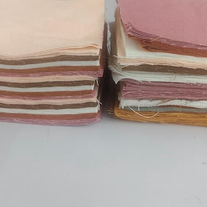 May include: Two stacks of fabric squares in shades of pink, brown, and cream. The fabric has visible edges, suggesting it's cotton or a similar material. The stacks are side by side on a white surface, possibly for a craft or sewing project.