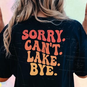 Puede incluir: Una camiseta negra con el texto "Sorry Can't Lake Bye" en una fuente retro y colorida.