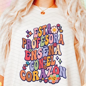 May include: A white t-shirt featuring a colourful graphic design that reads "Esta Profesora Enseña Con El Corazón" with flowers and stars.