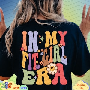 Può includere: Una maglietta nera con un design grafico colorato che dice "In My Fit Girl Era". La grafica è in stile retrò con fiori e un viso sorridente.