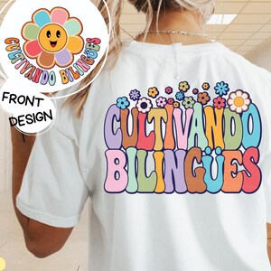 May include: White t-shirt featuring a colourful graphic design that reads "Cultivando Bilíngues" in a retro font. The design includes a flower with a smiling face and a border of smaller flowers.