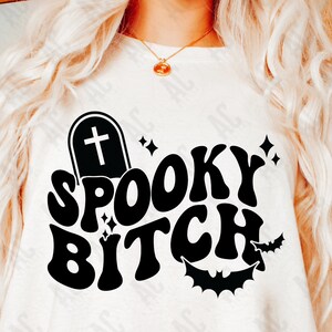 Puede incluir: Una camiseta blanca con un diseño gráfico negro que dice "Spooky Bitch" en una fuente retro. El gráfico también incluye una lápida con una cruz y murciélagos.