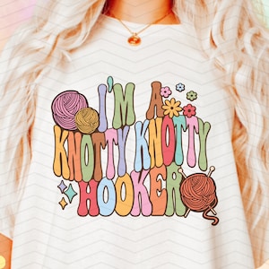 May include: A white t-shirt with the text "I'm a Knotty Knotty Hooker" in colorful, retro-style lettering. The design includes yarn balls and knitting needles.
