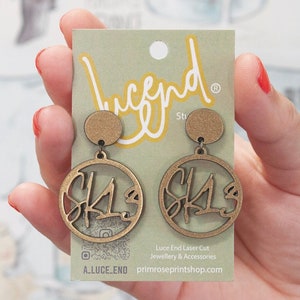 May include: Gold-coloured laser-cut earrings featuring the letters "SK13" within a circular design. The earrings are presented on a light green card with the "Lucend Studio" logo.