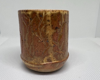May include: A handmade ceramic pot with a textured, earthy brown glaze. The cylindrical vessel features a detailed leaf pattern carved into the surface. The rim and base have a lighter, golden-brown hue, adding depth to the design.