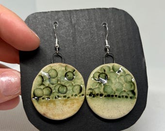 May include: A pair of ceramic earrings with a circular design. The top half of each earring features a textured pattern of green circles, while the bottom half is a light beige color. The earrings hang from silver-colored hooks.