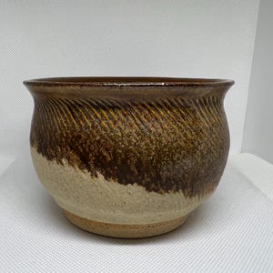 May include: A handcrafted ceramic bowl with a wide, rounded shape. The upper portion features a textured, diagonal pattern in shades of brown and gold, transitioning to a lighter beige base. The rim is a darker brown.