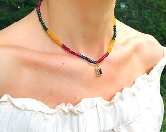 Puede incluir: Un collar de capas con cuentas verdes, amarillas, rojas y azules. El collar tiene un pequeño colgante con una piedra preciosa negra.