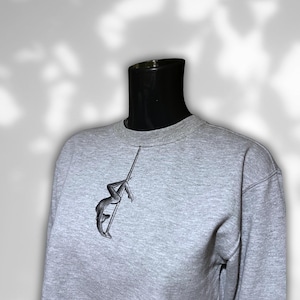 Könnte beinhalten: Hellgraues Sweatshirt mit Rundhalsausschnitt. Auf der linken Brust befindet sich ein schwarzes, gesticktes Design einer Person, die an einer Stange tanzt. Das Sweatshirt wird auf einer schwarzen Schaufensterpuppe präsentiert.
