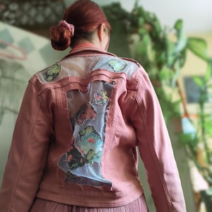 Può includere: Giacca di jeans rosa con toppe floreali e a farfalla sulla schiena. La giacca presenta un design invecchiato con inserti in tessuto trasparente. Le maniche sono lunghe e viene indossata sopra una gonna plissettata.