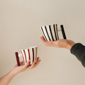 May include: Two ceramic mugs with striped designs. One mug is black and white, the other is red and white. Both mugs have a unique handle with a round knob.