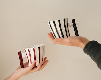May include: Two ceramic mugs with striped designs. One mug is black and white, the other is red and white. Both mugs have a unique handle with a round knob.