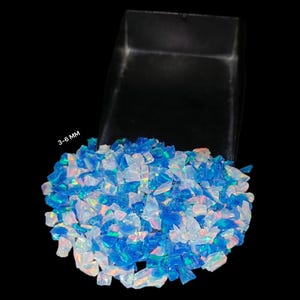 May include: A pile of blue and white opal chips, ranging from 3-6 mm in size, sits next to a black container. The opals have a shimmering, iridescent quality, with flashes of color. The background is black.