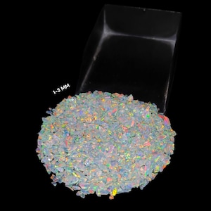 May include: A pile of iridescent opal chips, ranging in size from 1-3 mm, in a clear plastic tray against a black background.