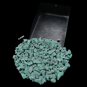 May include: A collection of small, teal-coloured stones, approximately 3-5 mm in size, spilling from a dark grey container. The stones are a vibrant green-blue hue, with a slightly rough texture. The background is black.