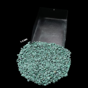 Puede incluir: Un montón de grava pequeña de color verde azulado, con un tamaño de 1-3 mm, se muestra saliendo de una pala negra sobre un fondo negro. La grava parece ser piedra triturada, posiblemente para paisajismo o fines decorativos.