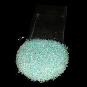 May include: A mound of pale blue crushed opal gemstones with iridescent specks, beside a black container. The text "0.5-1 MM" is visible. The gemstones are on a black background.