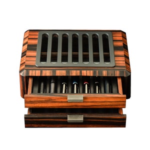 May include: A rectangular wooden pen storage box with a dark brown and black striped wood grain finish. The box features two pull-out drawers with metal handles and a top lid with a gray slotted panel that reveals the pens stored inside the case.