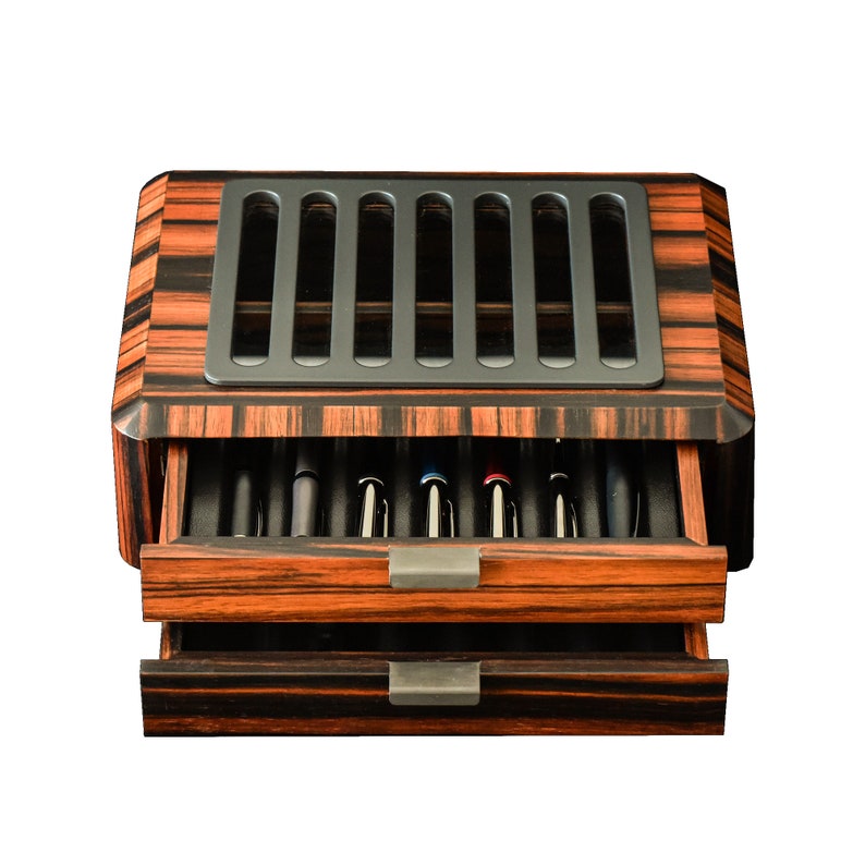 May include: A rectangular wooden pen storage box with a dark brown and black striped wood grain finish. The box features two pull-out drawers with metal handles and a top lid with a gray slotted panel that reveals the pens stored inside the case.