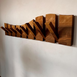 Könnte beinhalten: Ein Wandgarderobe aus Holz mit einer Reihe abgewinkelter Haken. Die Garderobe ist aus warm getöntem Holz gefertigt, wobei eine natürliche Maserung sichtbar ist. Die Haken sind zum Aufhängen von Mänteln, Hüten oder anderen Gegenständen konzipiert. Das Design ist modern und minimalistisch.
