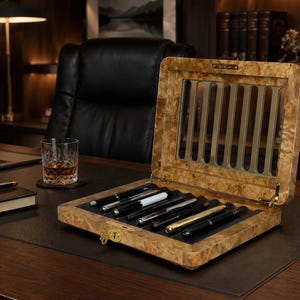 May include: A rectangular wooden display case with a burl wood finish and a hinged lid featuring a glass window. The interior contains six slots holding various fountain pens. The case has a gold-colored metal latch and a gold-colored brand plaque.