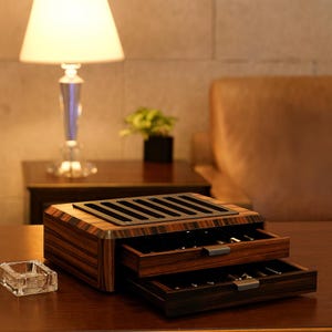 May include: A rectangular wooden storage box with a dark brown striped grain pattern, featuring two pull-out drawers with metallic handles and a black slotted top panel designed for organizing pens or similar items, resting on a flat surface.