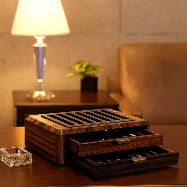 May include: A rectangular wooden storage box with a dark brown striped grain pattern, featuring two pull-out drawers with metallic handles and a black slotted top panel designed for organizing pens or similar items, resting on a flat surface.