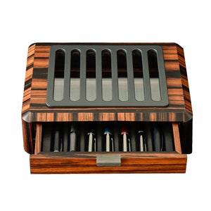 May include: A rectangular wooden pen display case with a dark brown and black striped wood grain finish. The top features a gray metal grate with seven vertical slots. A single pull-out drawer at the front contains seven pens arranged in individual slots.