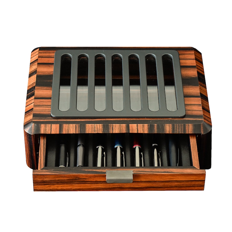 May include: A rectangular wooden pen display case with a dark brown and black striped wood grain finish. The top features a gray metal grate with seven vertical slots. A single pull-out drawer at the front contains seven pens arranged in individual slots.