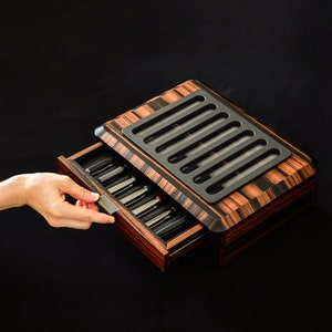 May include: A rectangular wooden pen storage box with a dark brown and black striped grain pattern. The box features a top panel with six vertical glass viewing slots and a pull-out drawer containing multiple black pens with silver clips.
