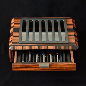 May include: A rectangular wooden pen display case featuring a dark brown and black striped wood grain finish. The top has a gray slotted viewing window, and a pull-out drawer at the front reveals several pens stored in individual black slots.