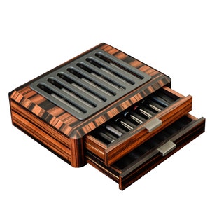 May include: A rectangular wooden storage box with a dark brown and black striped wood grain finish. The top features a gray slotted panel, and the front includes two pull-out drawers with metallic handles, each containing multiple black pens.