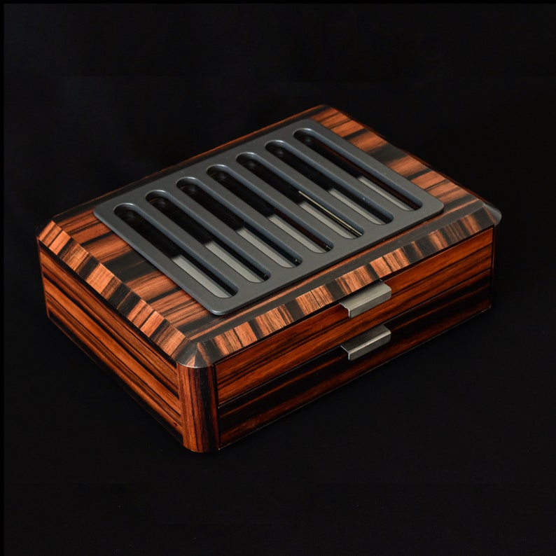 May include: A rectangular wooden storage box with a dark brown and black striped wood grain finish. The top features a matte gray panel with six vertical cutouts. Two drawers are stacked on the front, each with a simple, horizontal metal handle.
