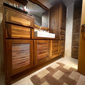 Può includere: Mobile da bagno in legno su misura con specchio coordinato. Il mobile presenta una combinazione di cassetti e ante a lamelle, che mostrano una ricca e calda venatura del legno. Un lavabo rettangolare bianco è integrato nel piano di lavoro. Un tappeto fantasia è sul pavimento.
