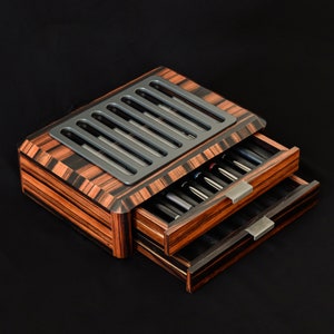 May include: A wooden storage box with a dark brown and black striped grain pattern, featuring two pull-out drawers with metallic handles and a top panel with a series of six vertical rectangular cutouts revealing internal contents.