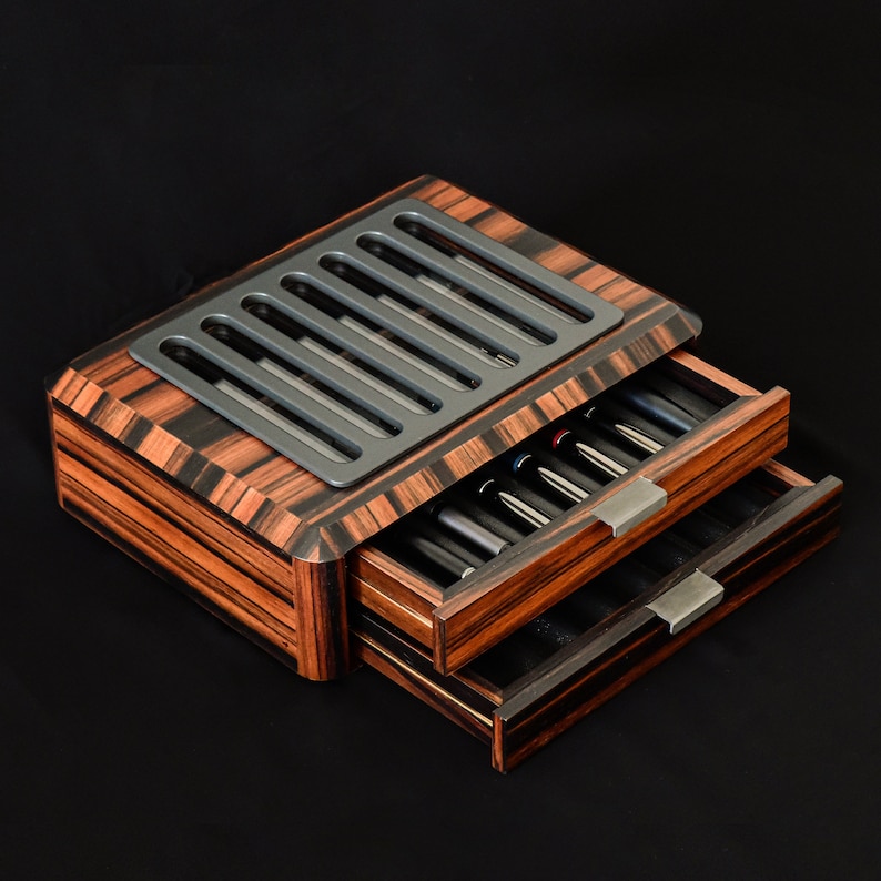 May include: A wooden storage box with a dark brown and black striped grain pattern, featuring two pull-out drawers with metallic handles and a top panel with a series of six vertical rectangular cutouts revealing internal contents.