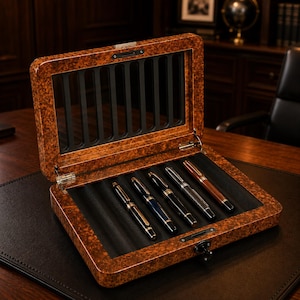 May include: A rectangular wooden display case with a burl wood finish and a hinged lid featuring a glass viewing window. The interior is lined with black material and contains five fountain pens arranged in individual slots within the box.