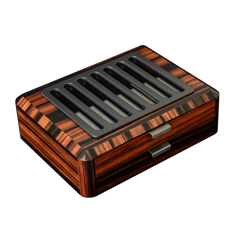 May include: A rectangular wooden box featuring a dark brown and black striped wood grain pattern. The top surface includes a matte black metal grate with five vertical slots. Two drawers are positioned on the front, each with a small metal handle.