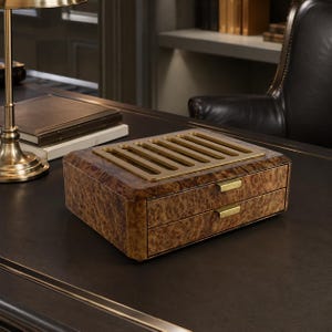May include: A rectangular wooden storage box with a burl wood finish, featuring two drawers with horizontal brass-toned metal handles and a slotted brass-toned metal plate on the top surface. The box has a smooth, polished appearance.