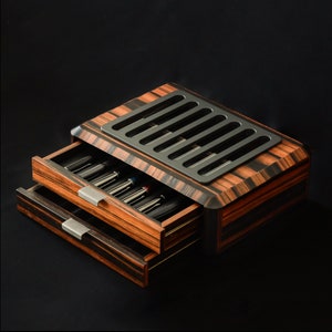 May include: A wooden storage box with a dark brown and black striped grain pattern featuring two pull-out drawers containing multiple pens. The top of the box has a rectangular black metal plate with six vertical slots for pen display.