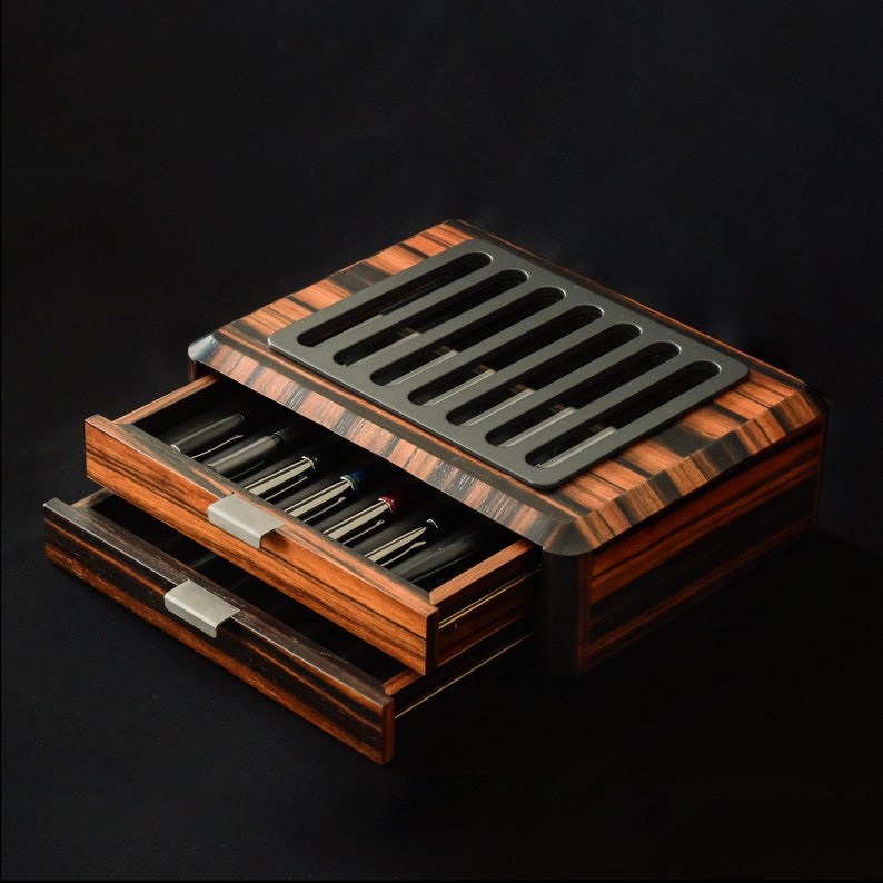 May include: A wooden storage box with a dark brown and black striped grain pattern featuring two pull-out drawers containing multiple pens. The top of the box has a rectangular black metal plate with six vertical slots for pen display.