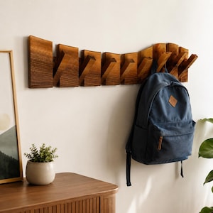 Puede incluir: Un perchero de pared de madera con ocho ganchos angulados. El perchero es de madera marrón oscuro y está montado en una pared blanca. Una mochila azul cuelga de uno de los ganchos. Una pequeña planta en maceta y un mueble de madera están en primer plano.