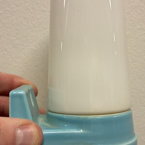 Könnte beinhalten: Eine Vintage-Wandleuchte. Die Leuchte hat einen zylindrischen, weißen Schirm auf einem hellblauen Sockel. Der Sockel hat ein geschwungenes Design und eine kleine Halterung. Die Leuchte wird von einer Hand gehalten.