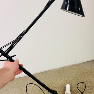 Könnte beinhalten: Eine schwarze, verstellbare Schreibtischlampe mit einem konischen Schirm und einem weißen Innenraum. Die Lampe hat einen flexiblen Arm und einen quadratischen Sockel. Eine weiße Glühbirne liegt neben der Lampe. Die Lampe steht auf einem beigen Teppich.