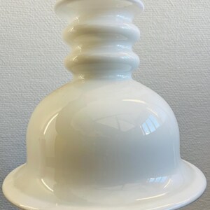 May include: A white ceramic pendant light fixture. The light has a bell-shaped shade with a flared rim and a cylindrical top with stacked, rounded ridges. The fixture is suspended by a thin, off-white cord.