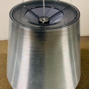 May include: A silver-colored, cone-shaped lamp with a dark gray top. The lamp has a textured, ridged surface and a central support with thin wires extending upwards. The lamp is designed for indoor use.