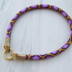 May include: A purple and gold beaded necklace with a snake-like pattern and gold metal clasps.