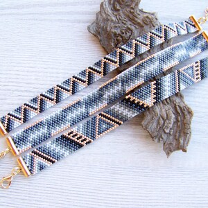 May include: Three beaded bracelets with geometric patterns in shades of gray, black, and white. The bracelets are connected with gold-toned clasps.