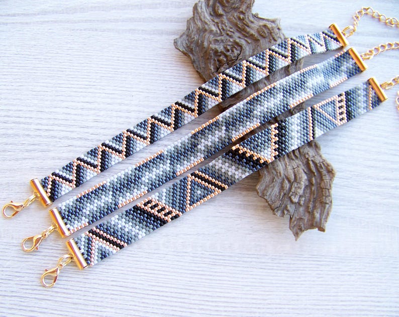 May include: Three beaded bracelets with geometric patterns in shades of gray, black, and white. The bracelets are connected with gold-toned clasps.